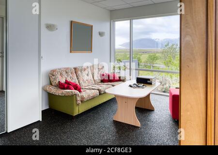 Neat clean cozy light living room with comfortable colorful sofa and red square pouf bedside wooden table on background of white walls and big window overlooking green garden and blurred foggy mountains Stock Photo