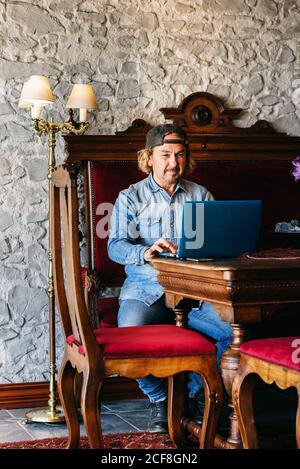 Adult male in denim clothes sitting at table with computer in retro style living room Stock Photo