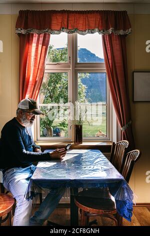 Side view of elderly bearded male sitting at table in cozy dining room near window and reading messages on mobile phone while relaxing during weekend Stock Photo