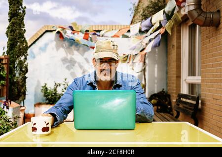Thoughtful elderly male in cap and denim shirt focusing on screen with interest while sitting at table with mug of tasty beverage and using netbook in yard of country house Stock Photo