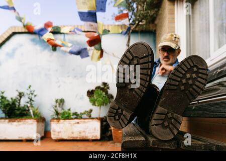 Low angle of calm pensive male pensioner in casual clothes and boots relaxing alone on wooden bench against green flowers growing in flowerbeds on terrace of cottage in suburb Stock Photo