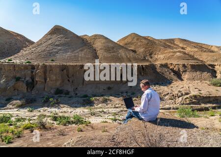 Traveler man working with laptop sitting on rocky mountain on beautiful scenic clif Stock Photo