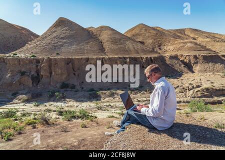 Traveler man working with laptop sitting on rocky mountain on beautiful scenic clif Stock Photo