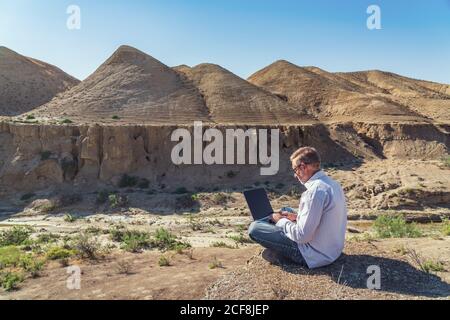 Traveler man working with laptop sitting on rocky mountain on beautiful scenic clif Stock Photo