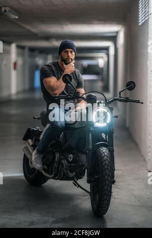 Full body bearded guy in casual clothes sitting on motorcycle and looking at camera in corridor of modern garage Stock Photo