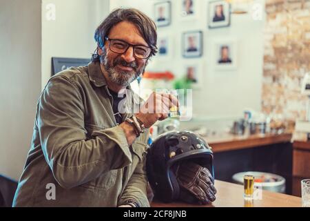 Happy bearded mature man with helmet smiling and looking at camera while drinking a alcohol shot in pub Stock Photo