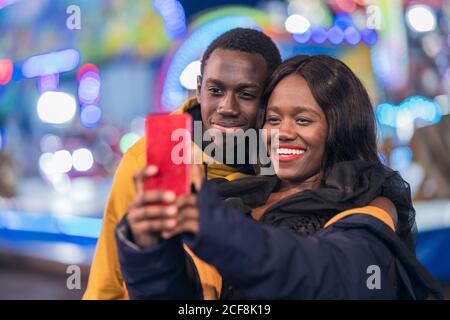 Cheerful African American man and Woman smiling and taking selfie during date on funfair at night Stock Photo