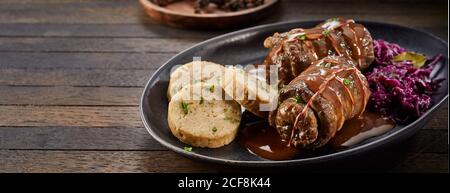 Double portion of beef roulades with bread dumplings and red cabbage served on a platter on a wooden table with copyspace in a panorama banner Stock Photo