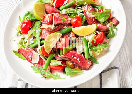 Green bean, cherry tomatoes and sliced beef steak salad with fresh arugula and shredded parmesan cheese served on a white plate with lime wedges on a Stock Photo