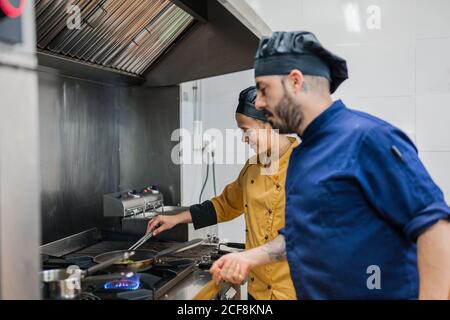 Side view of male chief chef watching young female assistant frying food while working together in professional kitchen Stock Photo