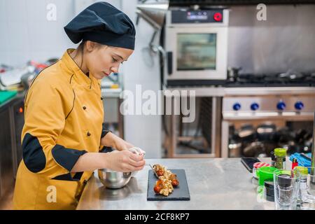 Side view of serious young female chef garnishing and finishing gourmet dish ready for serving while working at table in restaurant kitchen Stock Photo
