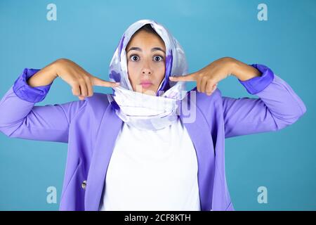 Young beautiful arab woman wearing islamic hijab over isolated blue background making fish face with lips, crazy and comical gesture. Stock Photo