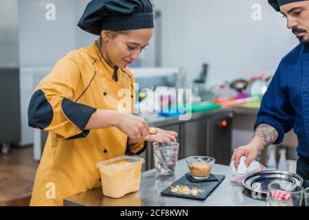 Male chef watching young female assistant with spoon in hand putting food on plate while working together in restaurant kitchen Stock Photo