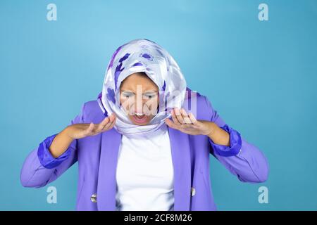 Young beautiful arab woman wearing islamic hijab over isolated blue background shouting and screaming loud down with hands on mouth Stock Photo