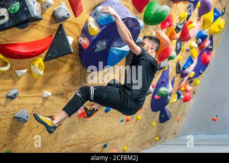 From bellow side view of strong male athlete in sportswear climbing on colorful wall during workout Stock Photo