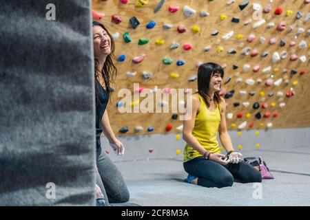 Happy female athletes sitting in lotus on knees on the floor while smiling and looking away resting during climbing training in gym Stock Photo