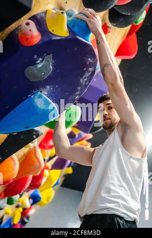 From below of young man in sportswear hanging on climbing wall and looking at camera during bouldering workout in gym Stock Photo