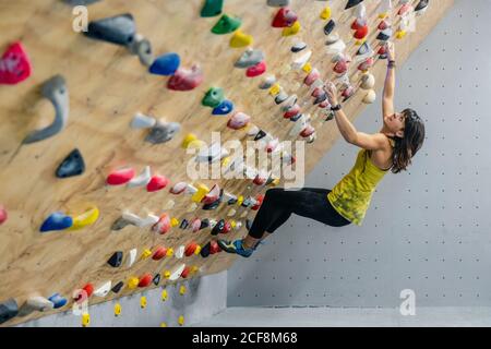 Side view from below of focused Woman in sportswear hanging on steep wall above mats in modern climbing center Stock Photo