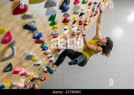 Side view from below of focused Woman in sportswear hanging on steep wall above mats in modern climbing center Stock Photo