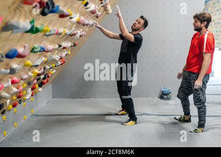 Side view of male athlete in sportswear climbing on grips of steep wall while male friend supporting with bag of talcum in gym Stock Photo