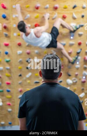 Back view of anonymous male athlete in sportswear watching for climber during workout on wall in gym Stock Photo