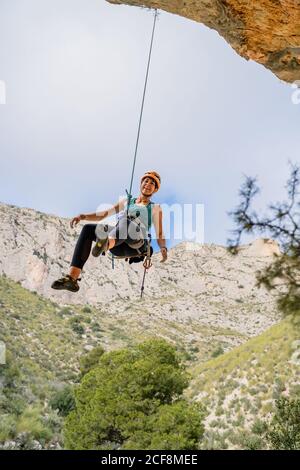 From below cheerful athletic female alpinist looking away while hanging on cliff under cloudy sky Stock Photo