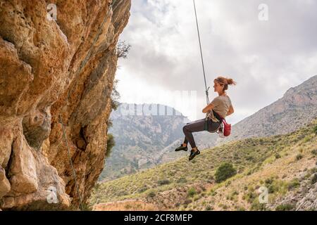 From below athletic female alpinist hanging on cliff under cloudy sky Stock Photo