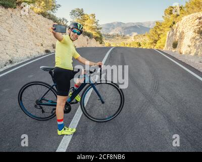 healthy man resting and taking selfie with smartphone while riding a bicycle on a mountain road in a sunny day Stock Photo