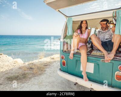 Tanned pretty Woman with mature man father sitting on open trunk of trailer at sandy seaside in bright day Stock Photo