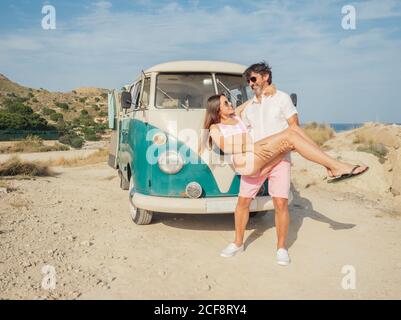 Side view of young pretty Woman in hands of stylish handsome man in sun glasses near old fashioned trailer at dusty shore Stock Photo