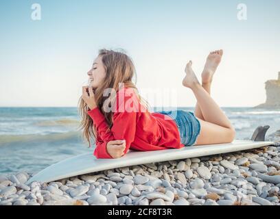 Side view of attractive smiling long haired Woman in red hoodie enjoying lying on surfboard on stony seaside Stock Photo
