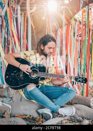 Bearded man sitting under decorated tree playing guitar Stock Photo