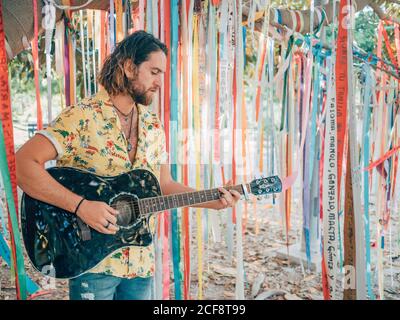 Bearded hipster man playing acoustic guitar under decorated tree on summer day Stock Photo