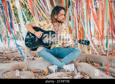 Bearded man sitting under decorated tree playing guitar Stock Photo