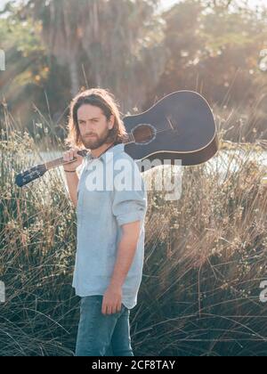 Pensive bearded hipster man walking in jungle with guitar on shoulder looking at camera Stock Photo