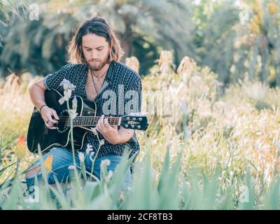 Pensive bearded hipster man sitting in meadow playing guitar Stock Photo