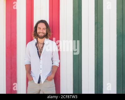Confident bearded man in casual outfit holding hands in pockets and cheerfully smiling while standing against striped wall on resort Stock Photo