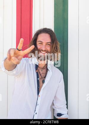 Confident and happy bearded man in casual outfit with victory sign and cheerfully smiling while standing against striped wall on resort Stock Photo