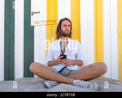 Confident bearded man in casual outfit using smartphone while standing against striped wall on resort Stock Photo