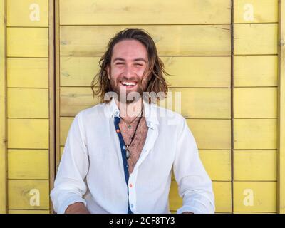 Confident bearded man in casual outfit and cheerfully smiling while sitting against blue wall on resort Stock Photo