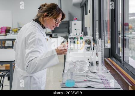 Side view of mature female scientist with clipboard examining glassware while working in modern chemistry laboratory Stock Photo