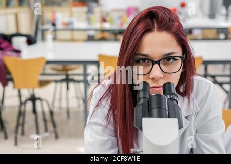 Young Woman withe red hair looking at camera while using microscope on blurred background of lab Stock Photo
