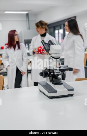 Modern microscope placed on table against female scientist and young students in contemporary laboratory Stock Photo
