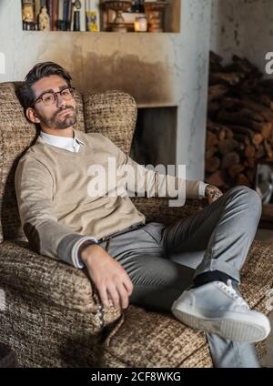 Serious confident man in stylish casual clothes with glasses looking at camera and contemplating while sitting in soft brown armchair against vintage interior Stock Photo