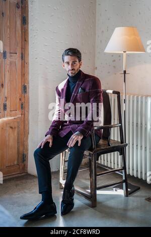 Serious adult man in elegant clothes looking at camera while sitting on aged wooden armchair beside white radiator and vintage floor lamp against spotted white wall in country house Stock Photo