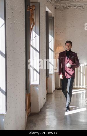 Bossy confident executive man in elegant burgundy jacket and black pants and shoes walking along light spacious corridor against vintage interior in office building Stock Photo
