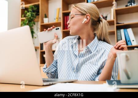 Image of happy woman using earphones and singing while working with laptop at table in office Stock Photo
