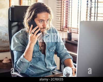Concentrated brutal man in casual shirt communicating on smartphone while using computer in home office Stock Photo
