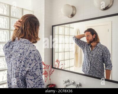 Back view of cool adult man fixing hair in standing in light bathroom in front of mirror Stock Photo