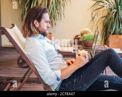 Side view of brutal bearded man eating apple and using laptop while relaxing on deckchair on balcony at home Stock Photo